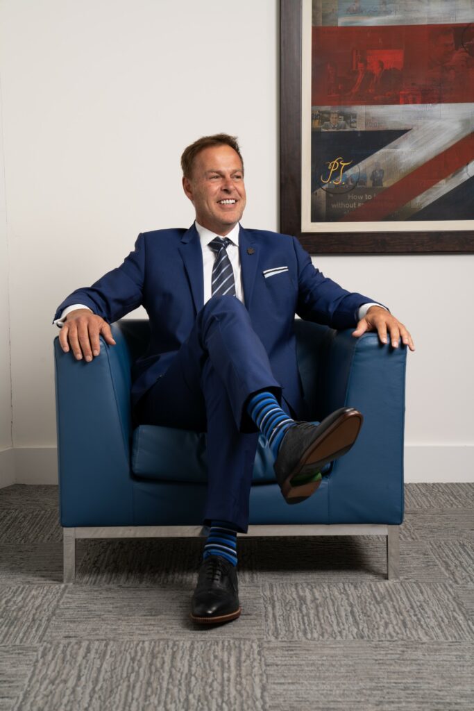 Image of PEter Jones sat in a blue fabric arm chair, crossed legs, smiling in an office without a desk wearing a blue suit and tie with a white shirt